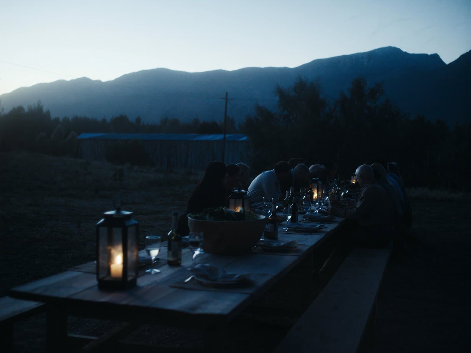 A long plank table set for dinner outdoors on a Western ranch at blue hour, lanterns lit.
